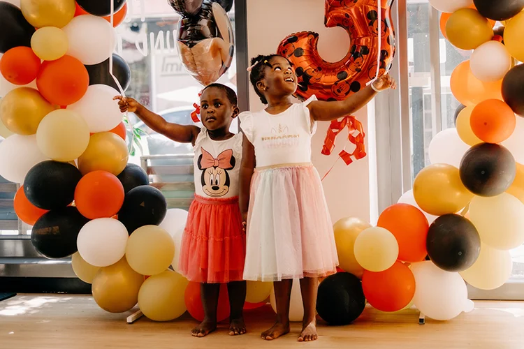 Zwei fröhliche junge Mädchen halten Luftballons in den Händen und stehen vor einem Luftballon-Bogen auf der individuell geplanten Geburtstagsparty.
